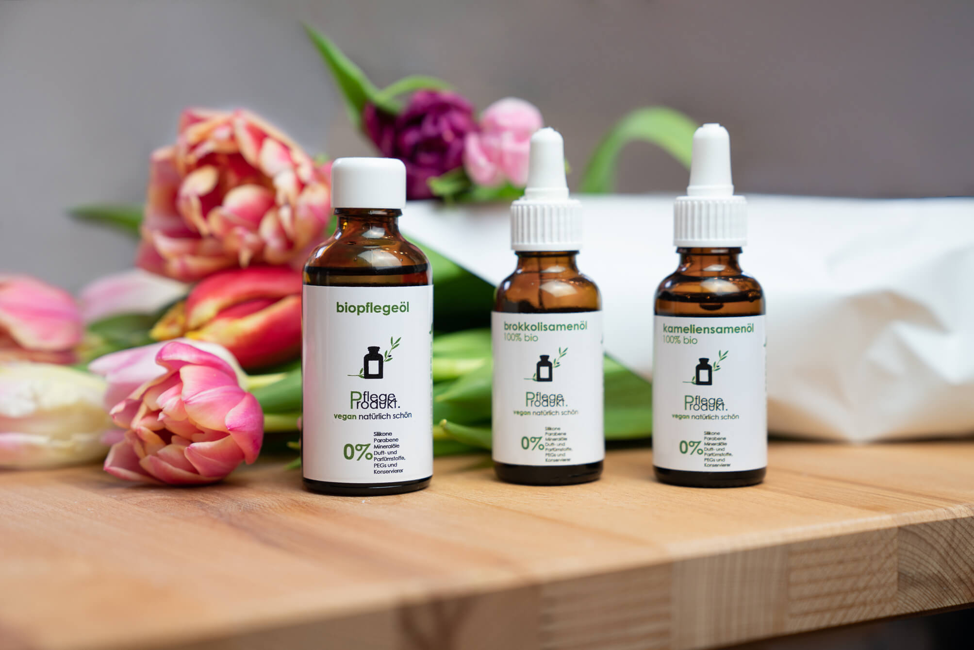 Drei Pflegeöle von Pflegeprodukt: Biopflegeöl, Bio Brokkolisamenöl, Bio Kameliensamenöl auf einem Holzbrett mit bunten Tulpen im Hintergrund.