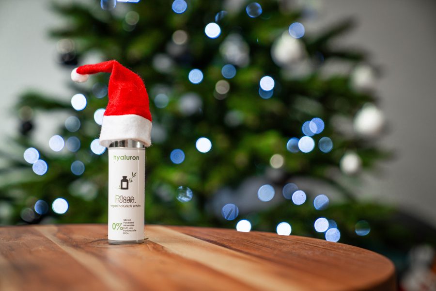 Hyaluron-Serum von Pflegeprodukt steht auf einem Holzbrett. Eine kleine rote Weihnachtsmann-Mütze steckt auf dem Behälter. Im Hintergrund ist ein Tannenbaum zu sehen.